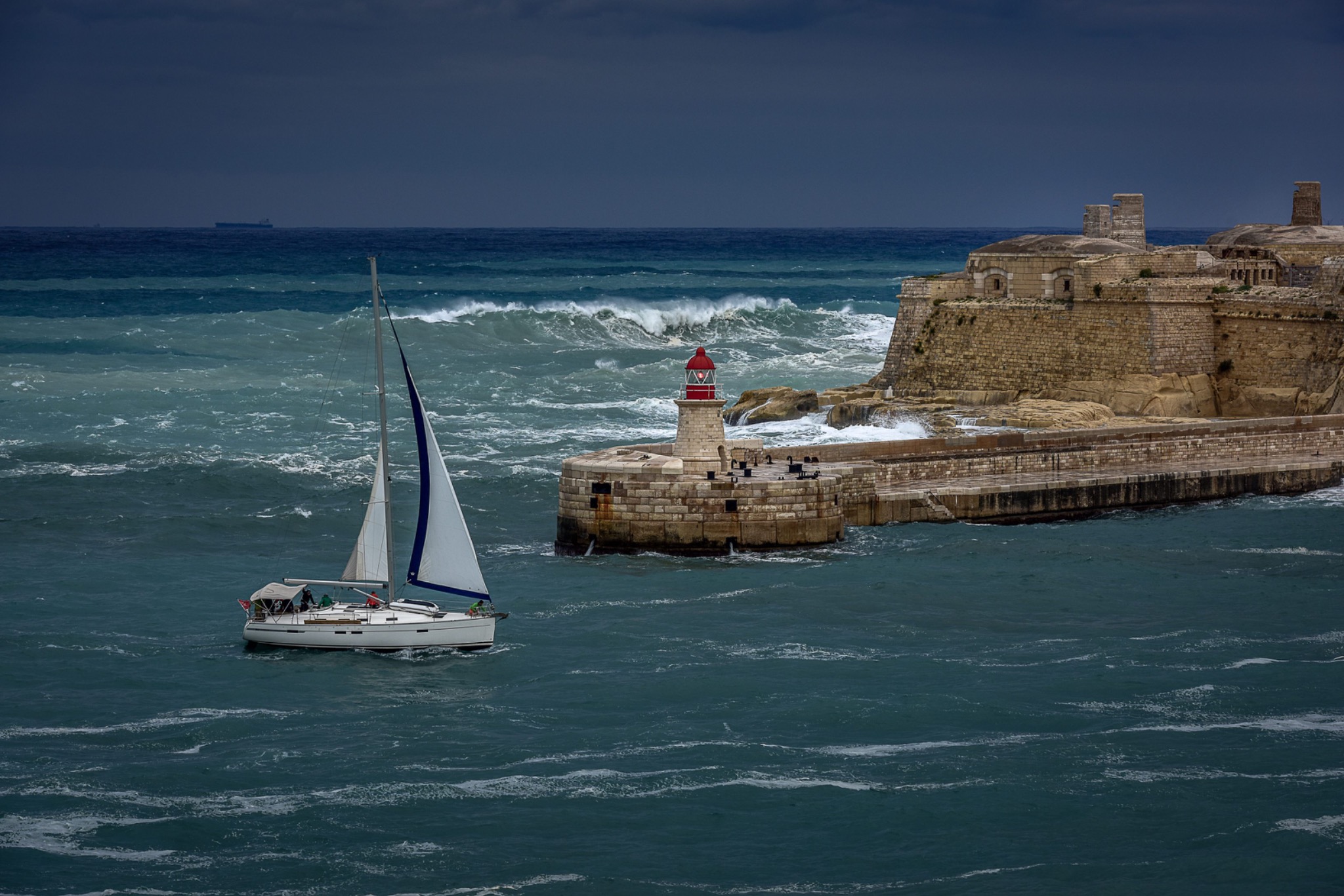 A sailing boat passing historic Maltese harbour fortifications and a red lighthouse.